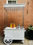 White mobile bar cart with drinks and champagne branding against a brick wall.