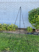 A black collapsible lightweight easel positioned on grass against a brick wall with shrubbery to the side.