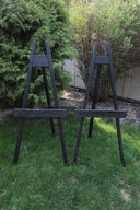 Two black wooden easels standing on grass with bushes in the background.