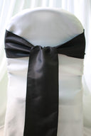 A black satin chair sash tied in a bow on a white chair cover, providing a sleek and elegant appearance.