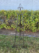 A black iron easel standing on grass with a hedge in the background.