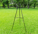 A black expandable easel set up on a grassy field with trees in the background.