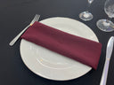 A burgundy polyester napkin is placed on a white plate on a black tablecloth, with a fork on the left side and a knife on the right side of the plate.