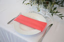 A coral colored polyester napkin on a white plate, with a fork on the left side and a knife on the right side of the plate, placed on a white tablecloth.
