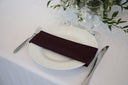 A eggplant purple polyester napkin is placed on a white plate on a table, with cutlery on either side and a glass vase with greenery in the background.