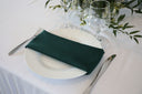 A hunter green polyester napkin is placed on a white plate, set on a table with cutlery and a white tablecloth.