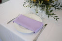 A lavender colored polyester napkin is placed on a white plate on a table with a floral arrangement in the background.