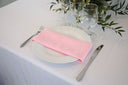 A light pink polyester napkin is placed on a white plate on a table with a white tablecloth. The napkin is neatly folded, and there are glasses and greenery in the background.
