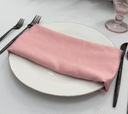 Pink napkin on a white plate with black cutlery and a glass in the background.