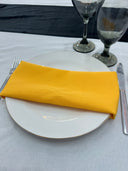 A marigold yellow polyester napkin is placed on a white plate on a table with a black border and cutlery visible.