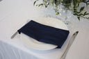 A navy polyester napkin is placed on a white plate, which is itself placed on a white tablecloth. The napkin is neatly folded, and there are silverware items on either side of the plate.