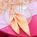 A peach satin napkin is displayed on a plate with a fork, knife, and spoon placed beside it, set against a pink tablecloth.