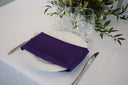 A purple polyester napkin is placed on a white plate on a table, with a glass and a floral arrangement in the background.