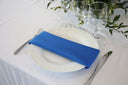 A royal blue polyester napkin is placed on a white plate on a table with glassware and greenery in the background.