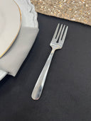 A silver dessert fork placed on a dark surface with a plate in the background.