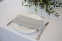 A silver polyester napkin is placed on a white plate on a table with cutlery and a floral arrangement in the background.
