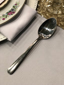 A silver spoon placed on a table with a decorative plate and a cloth in the background.