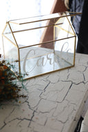 A gold glass card box with the word 'Cards' written on it, placed on a table with a decorative item in the background.