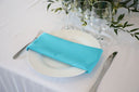 A turquoise polyester napkin is placed on a white plate on a table, with a glass and a fork/knife in the background.