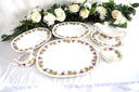 A vintage ceramic serving set with gold trim featuring grape and leaf designs, displayed on a table with a floral arrangement.