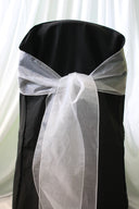 A white organza chair sash tied in a bow on a black chair cover, with a sheer white curtain in the background.