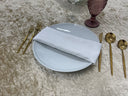 A white satin napkin is placed on a white plate, surrounded by gold-colored cutlery on a textured grey surface.
