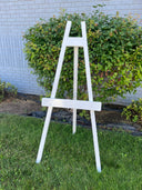 A white wooden easel standing on grass against a brick wall with greenery in the background.