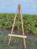 A wooden easel outdoors with a blue wall in the background and greenery around it. It is set up on a grassy area and appears to be sturdy with a horizontal board at the base.