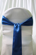 A blue satin chair sash tied around a white chair cover, with a focus on the sash which has a slight sheen due to its satin finish.