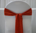 A burnt orange satin chair sash tied in a bow on a white chair cover.