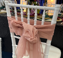 A dusty rose colored chiffon chair sash tied around a white chair post, with a decorative bow at the back.