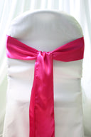 A fuchsia satin chair sash tied in a bow on a white chair cover, with a blurred background.