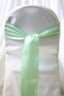 A mint satin chair sash tied in a bow on a white chair cover, with a blurred background.