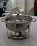 Stainless steel chafing dish on a table with chairs and a tablecloth in the background